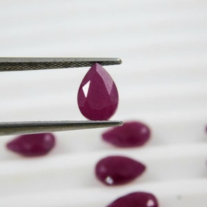 May include: A group of pear-shaped ruby gemstones, one of which is held by tweezers. The rubies are a deep red color and have a smooth, polished finish.