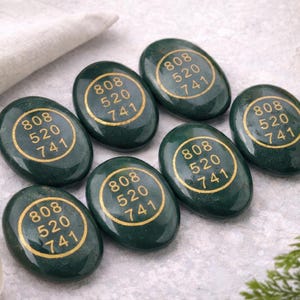 Puede incluir: Siete piedras ovaladas de color verde oscuro con letras doradas. Cada piedra tiene los números "808 520 741" en un círculo dorado. Están dispuestas sobre una superficie gris claro, con un paño blanco y vegetación.