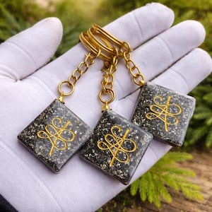 May include: Three square keychains with gold-colored hardware. Each keychain features a dark gray, speckled square pendant with a gold-colored, stylized design. The keychains are connected to a gold-colored key ring.