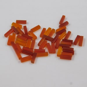 May include: A scattering of small, rectangular, translucent beads in shades of orange and red. The beads are spread across a white surface, with varying degrees of transparency and colour intensity. These beads could be used for jewellery making or other craft projects.