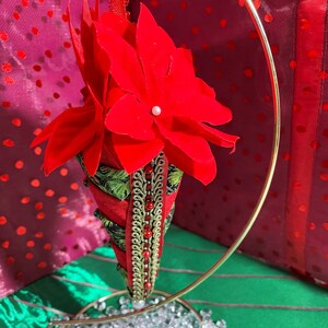 Puede incluir: Adorno navideño de flor de pascua roja con soporte metálico dorado. El adorno en forma de cono presenta una flor de terciopelo rojo y adornos verdes y dorados. Hay cuentas de vidrio transparente en la base del soporte. El fondo tiene un patrón de puntos rosas y rojos.