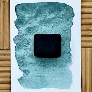 May include: A square, black watercolor paint pan sits on a teal watercolor swatch. The swatch is painted on white paper, with a textured appearance. The background is a bamboo surface.