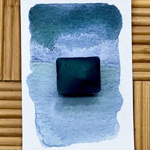 May include: A square, dark teal watercolor paint block sits on a watercolor swatch with shades of blue and teal. The background is a light blue and teal wash. The background is a bamboo surface.