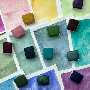 May include: A collection of rectangular watercolor paint pans in various solid colors, including green, blue, pink, and purple, arranged over a background of hand-painted watercolor swatches that display corresponding color gradients and textures.