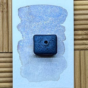 May include: A square, dark blue watercolor paint pan with a small hole in the center sits on a white card with a light blue watercolor swatch. The swatch has a shimmering, glittery effect.