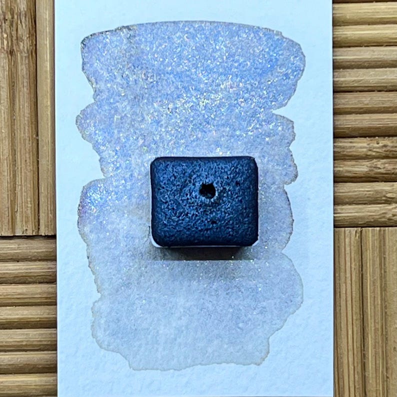 May include: A square, dark blue watercolor paint pan with a small hole in the center sits on a white card with a light blue watercolor swatch. The swatch has a shimmering, glittery effect.