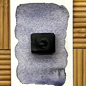 May include: A square, black watercolor paint pan sits on a white paper with a blue-grey watercolor swatch. The paper has a torn edge. The background is a bamboo mat.