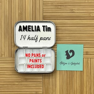 May include: A silver metal watercolor tin with the text "AMELIA Tin 14 half pans" on the lid. The tin is open and shows 14 empty half pans. The text "NO PANS or PAINTS INCLUDED" is printed on the inside of the tin. A teal blue square with a black squirrel silhouette and the text "Addison & Sedgwick" is in the background.