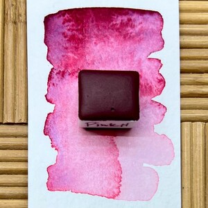 Op de afbeelding: Een rechthoekig aquarelverf napje in een diepe donkere kastanjebruine tint, gelabeld met de handgeschreven tekst Pink H, geplaatst op een wit papierstaal met een verloop van levendige magenta en roze aquarelverfstreken en texturen.