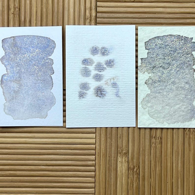 May include: Three watercolor swatches on white paper, showcasing shimmering, light blue and silver hues. The swatches are arranged on a bamboo surface, with the paper textures and color variations clearly visible. The artwork is a study of color and texture.