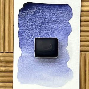 May include: A square, dark blue watercolor paint pan sits on a white paper with a blue wash. The paint pan is in a silver metal casing. The background is a light brown, textured surface.