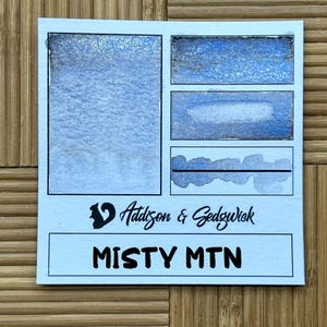 May include: A light blue watercolor paint swatch card with the text "Addison & Sedgwick" and "MISTY MTN". The card features several rectangular paint samples in shades of blue and silver, showcasing the paint's color and texture.