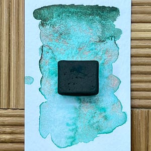 May include: A square, dark gray watercolor paint block sits on a watercolor swatch of teal and green hues. The background is a light blue and green watercolor wash with a textured appearance. The background is on a white paper.