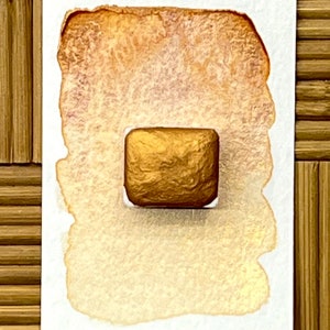 May include: A square, gold-colored watercolor paint pan sits on a white surface, surrounded by a light brown watercolor wash. The paint pan has a textured surface. The background is a bamboo mat.