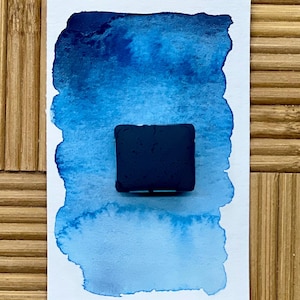 May include: A dark navy blue watercolor paint block sits on a white paper with a blue watercolor wash. The wash transitions from a deep navy to a light blue. The background is a bamboo surface.