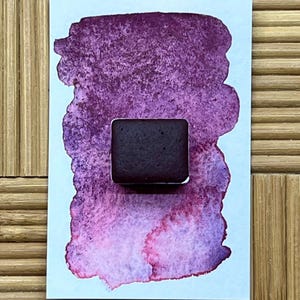 May include: A square, dark burgundy watercolor paint pan sits on a light blue paper with a textured, blended watercolor swatch in shades of purple and pink. The background is a light brown, textured surface.