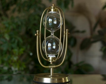 Può includere: Una clessidra dorata con un supporto decorativo. La clessidra ha due bulbi di vetro collegati da un collo stretto. Il supporto ha una base rotonda e un design curvo.
