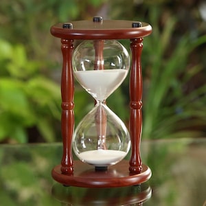 May include: A wooden hourglass with a brown base and clear glass bulbs. White sand flows through the hourglass.