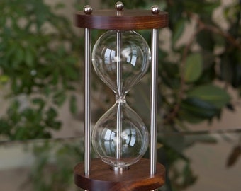 May include: A wooden hourglass with a silver frame and glass bulbs. The hourglass is sitting on a table with a reflective surface.