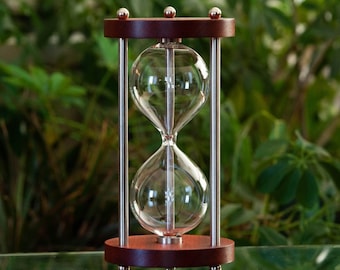 May include: An hourglass with a dark wood frame and base, featuring clear glass bulbs connected by a narrow neck. The hourglass is supported by four silver metal rods and topped with three silver spheres. The background is a blurred green foliage.