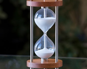 May include: A wooden hourglass with a silver frame and white sand. The hourglass is on a glass surface.