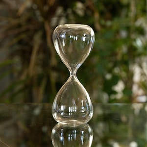 Puede incluir: Un reloj de arena de cristal transparente con una parte superior e inferior bulbosas, conectadas por un cuello estrecho. El reloj de arena está sobre una superficie reflectante, con un fondo verde borroso.
