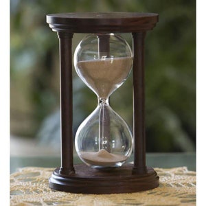 Puede incluir: Un reloj de arena de madera marrón oscuro con un bulbo de vidrio transparente lleno de arena. La arena fluye del bulbo superior al bulbo inferior.