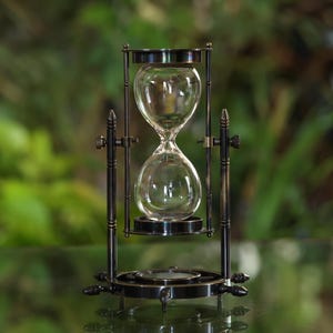 May include: A brass hourglass with a clear glass bulb and a stand with a compass design. The hourglass is on a reflective surface.