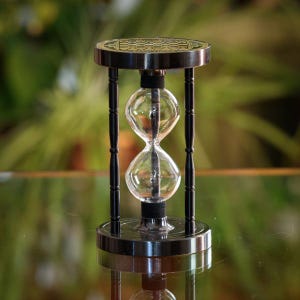 May include: An hourglass with a clear glass center and black metal frame. The top and bottom are circular with a decorative pattern. The hourglass is supported by four black pillars.