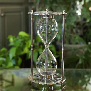May include: A silver hourglass with a clear glass bulb. The hourglass is on a silver base with Roman numeral markings.