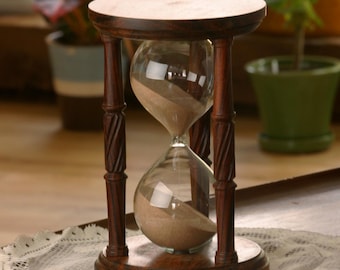 May include: A wooden hourglass with a dark brown finish. The hourglass has a clear glass bulb with sand inside. The sand is a light brown color. The hourglass is sitting on a white lace tablecloth.