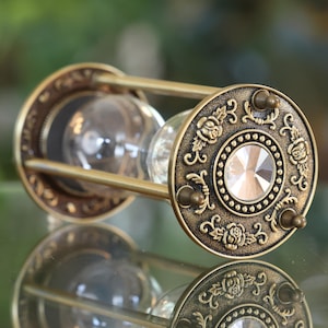 May include: An ornate brass hourglass with a clear glass bulb and a decorative crystal on the bottom. The hourglass is decorated with a floral pattern and has a vintage look.