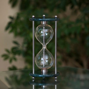 May include: A decorative hourglass featuring a clear glass bulb, supported by a green marble base and top, connected by silver metal rods. The background is a blurred green, creating a classic and elegant aesthetic.