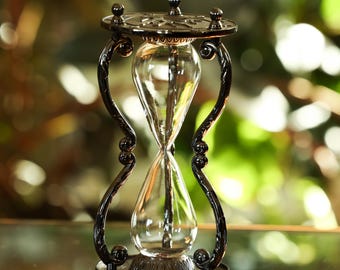 Può includere: Una clessidra con un centro in vetro trasparente e una cornice in metallo scuro. La cornice presenta supporti curvi decorativi e una base circolare. La parte superiore e inferiore hanno dettagli ornati. Per misurare il tempo.