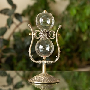 May include: A gold-toned hourglass with an ornate metal stand. The hourglass has two clear glass bulbs connected by a narrow neck. The stand has a decorative base with intricate details.