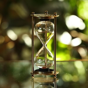 Könnte beinhalten: Eine Sanduhr aus Messing mit einer durchsichtigen Glasbirne. Die Sanduhr steht auf einer Glasfläche mit einem verschwommenen grünen Hintergrund.