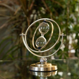 May include: A decorative hourglass with a silver-colored metal frame and base. The hourglass features a clear glass bulb filled with sand, set within a circular frame. The design is elegant and modern, suitable for home decor.