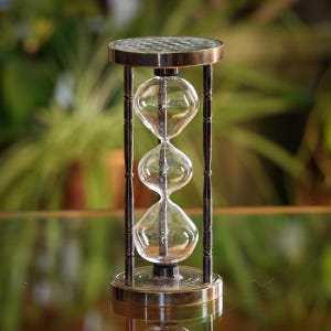 May include: An hourglass with a brass-colored frame and base, holding clear glass bulbs. The top and bottom are circular, with a patterned design. The hourglass is set against a blurred green background.