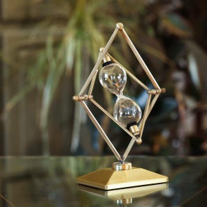 May include: A gold-toned hourglass with a geometric metal frame. The hourglass is suspended within the frame and rotates as the sand flows.