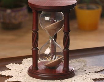 May include: A wooden hourglass with a dark brown finish. The hourglass has a round base and a cylindrical top. The sand is a light brown color. The hourglass is sitting on a white crocheted doily.