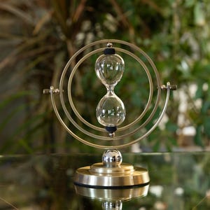 May include: An hourglass with a clear glass bulb, held within a gold-colored metal frame. The hourglass is supported by a gold-colored base. The background is blurred, with green foliage visible.