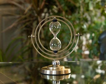 May include: An hourglass with a clear glass bulb, held within a gold-colored metal frame. The hourglass is supported by a gold-colored base. The background is blurred, with green foliage visible.