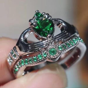 May include: A silver ring with two interlocking hands, a large green emerald gemstone, and smaller green emerald stones around the band.
