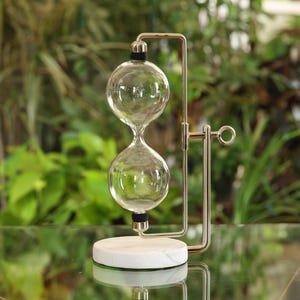 May include: A gold metal hourglass stand with a white marble base. The hourglass is made of clear glass and has two bulbs connected by a narrow neck.