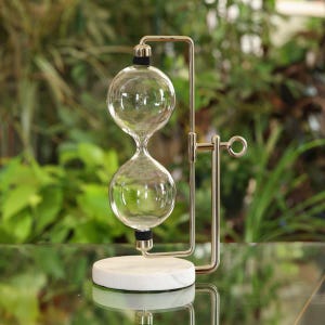 May include: A gold metal hourglass with a white marble base. The hourglass is made of clear glass and has a round shape. The hourglass is on a table with a green background.