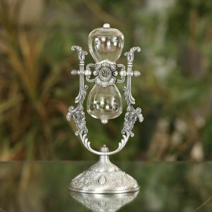 May include: A silver hourglass with an ornate stand, featuring floral designs. The hourglass is filled with clear sand and is suspended between two decorative arms.