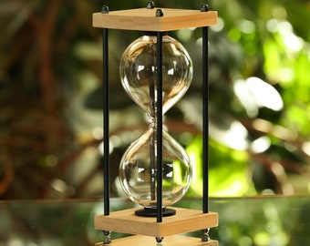 May include: A wooden hourglass with a black metal frame and a clear glass bulb. The hourglass is sitting on a table with a green background.