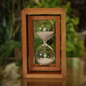 May include: A wooden hourglass with a clear glass bulb filled with white sand. The hourglass is encased in a rectangular wooden frame, with a second frame inside. The background is blurred, with green foliage.