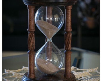 May include: A wooden hourglass with a dark brown finish and a glass bulb filled with sand. The hourglass is sitting on a white lace tablecloth.