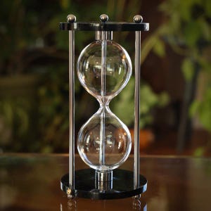 Op de afbeelding: Een zandloper met een heldere glazen bol en een zwarte basis en bovenkant. De zandloper wordt ondersteund door vier zilveren metalen staven. De achtergrond is wazig, met groen gebladerte zichtbaar.